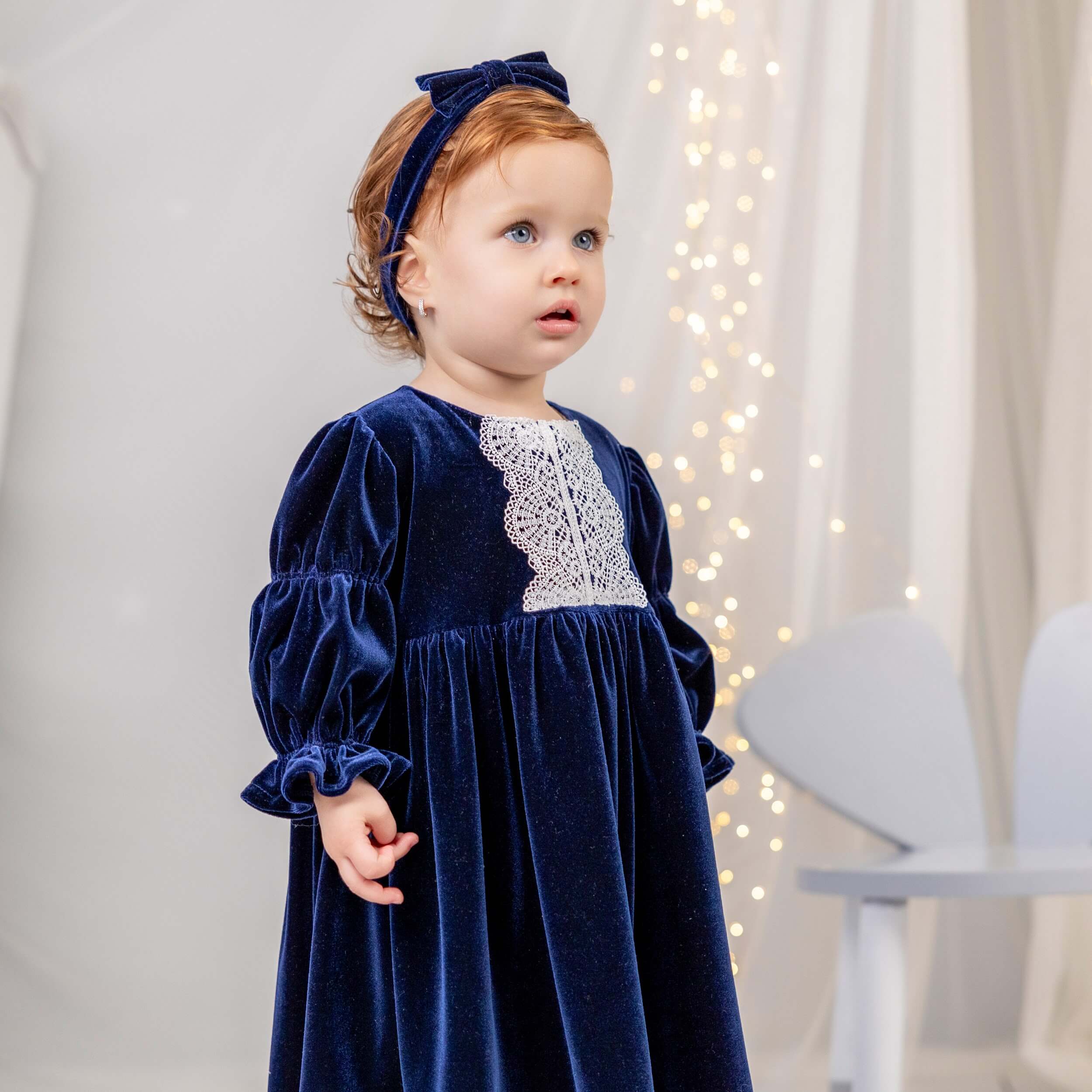 Child wearing a blue velvet dress with lace details in a softly lit room.