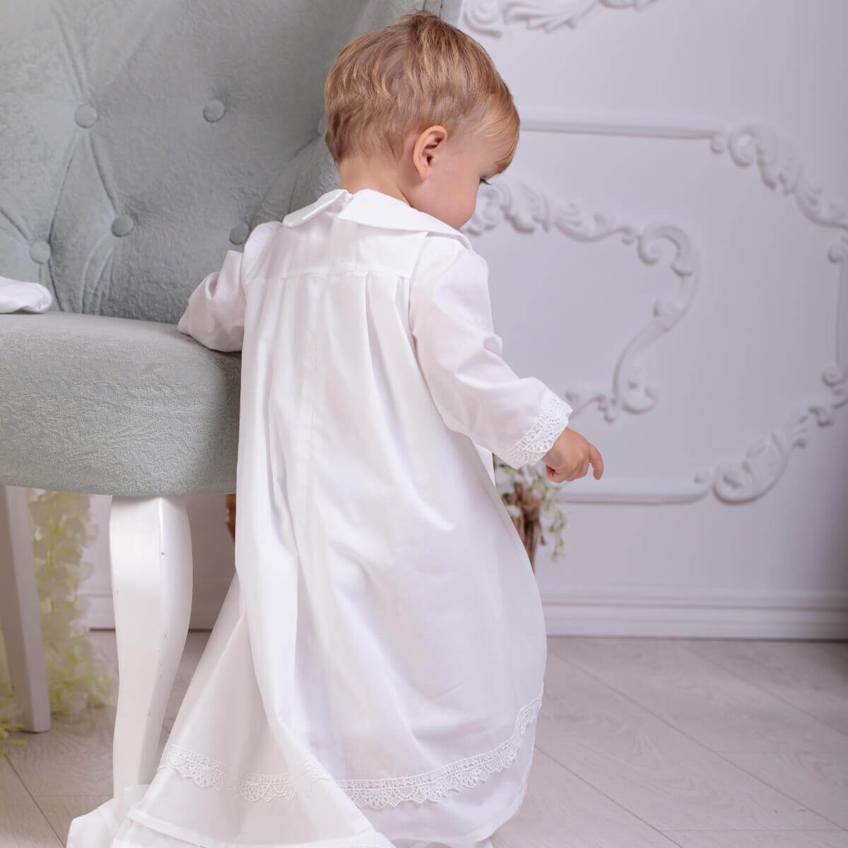 Child in a christening long gown standing next to a gray chair against a decorative white wall.