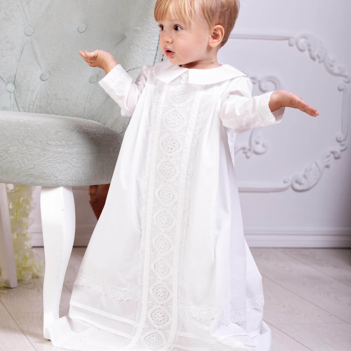 Child wearing a christening long gown standing in a room with a light-colored floor and wall.