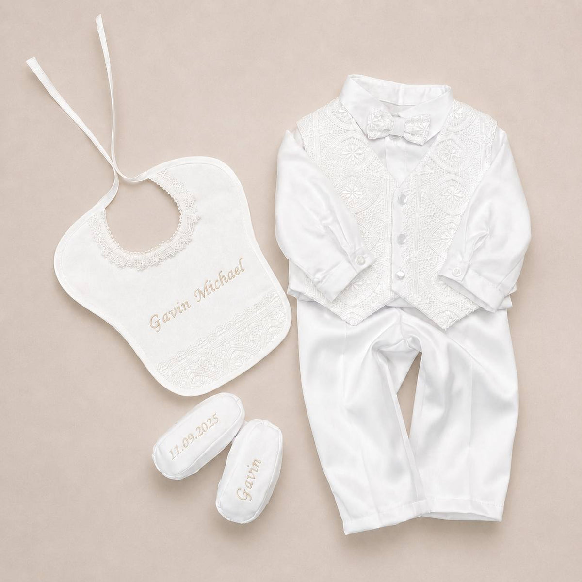 A baby formal outfit set laid flat on a light beige background, including a white satin long-sleeve shirt with lace chest panel and bow tie, a matching bib with lace trim and embroidered name, and two white booties with embroidered name and date.