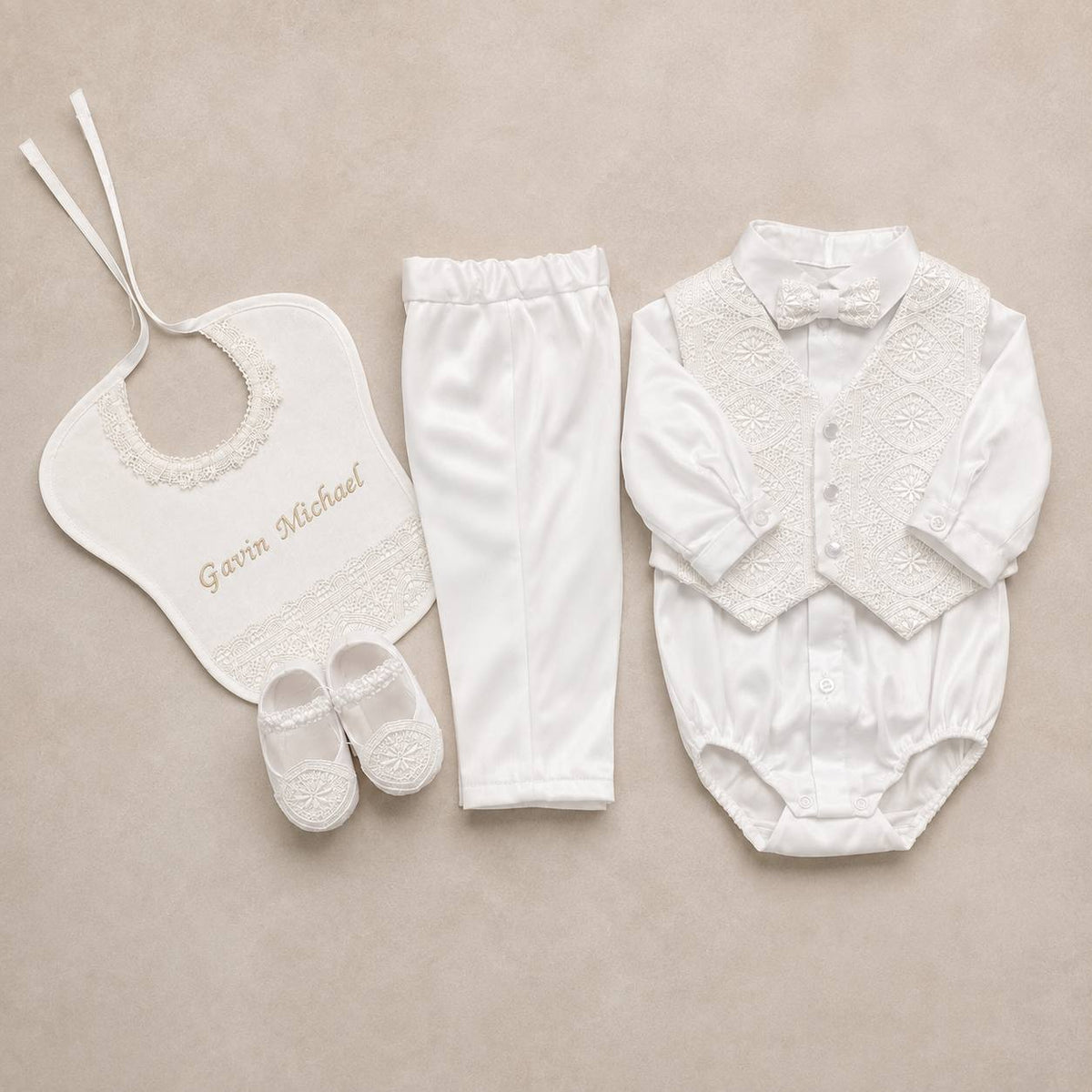 A white baby boy's formal outfit set laid flat on a beige background, including a long-sleeve lace-trim bodysuit with bow tie, white pants, a white bib with embroidered name 'Gavin Michael', and white booties.