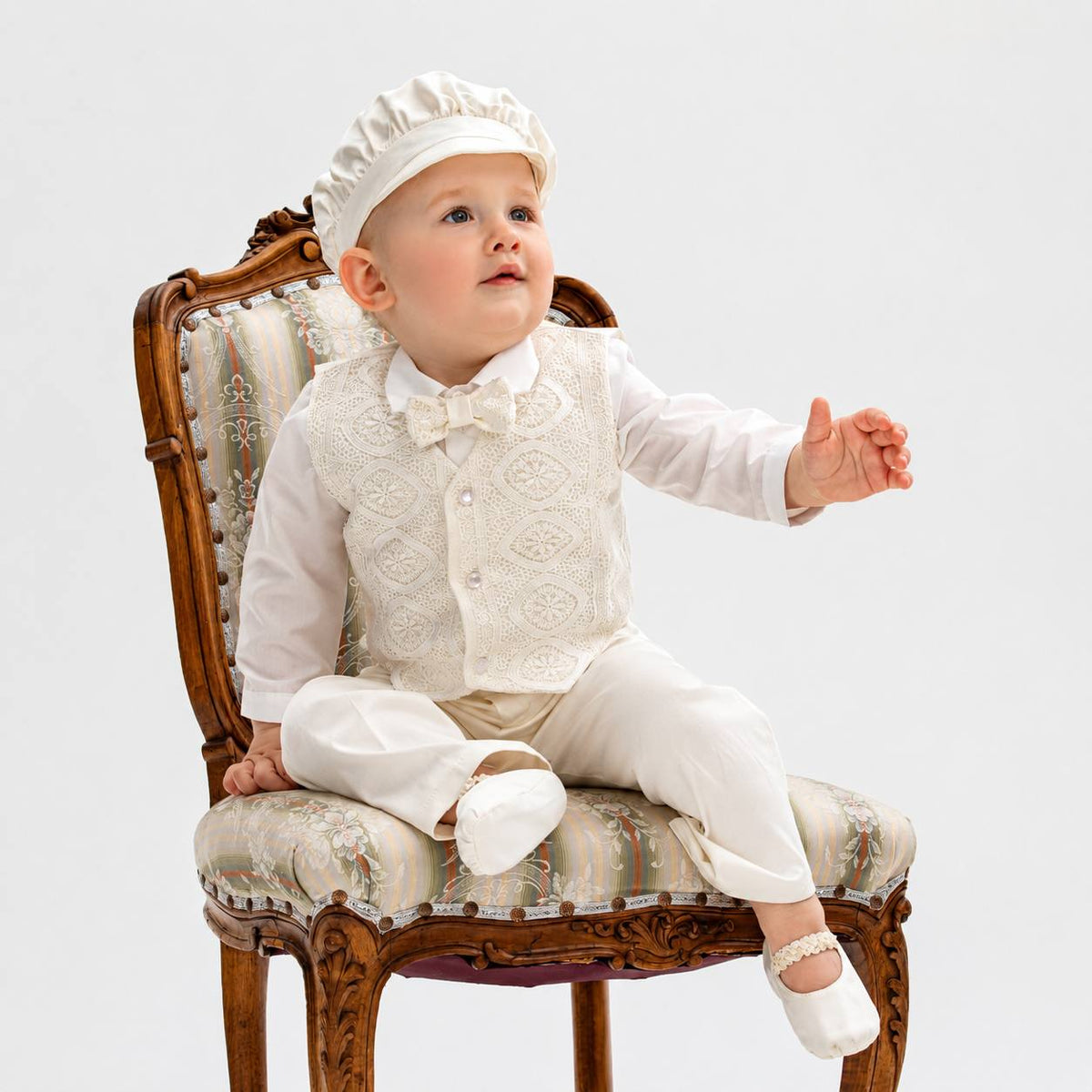 A baby wearing an elegant cream-colored formal outfit with a lace vest, matching bow tie, trousers, and a ruffled hat, sitting on an ornate wooden chair against a plain white background.