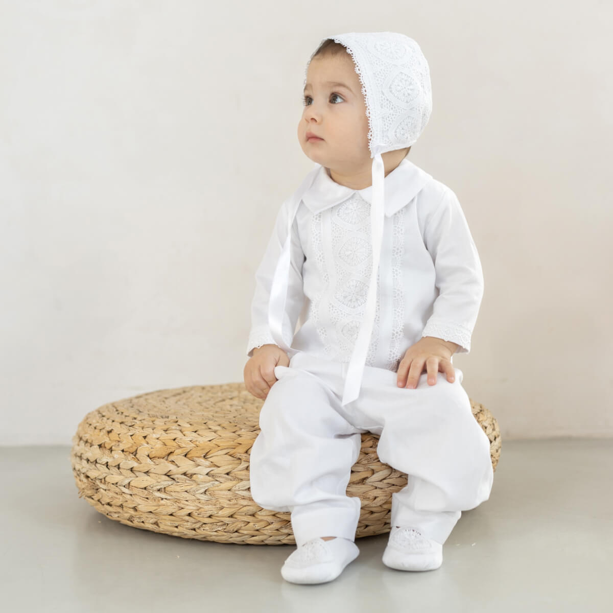 Baby in a Baby Boy Christening Suit  with a bonnet sitting on a woven stool against a plain background