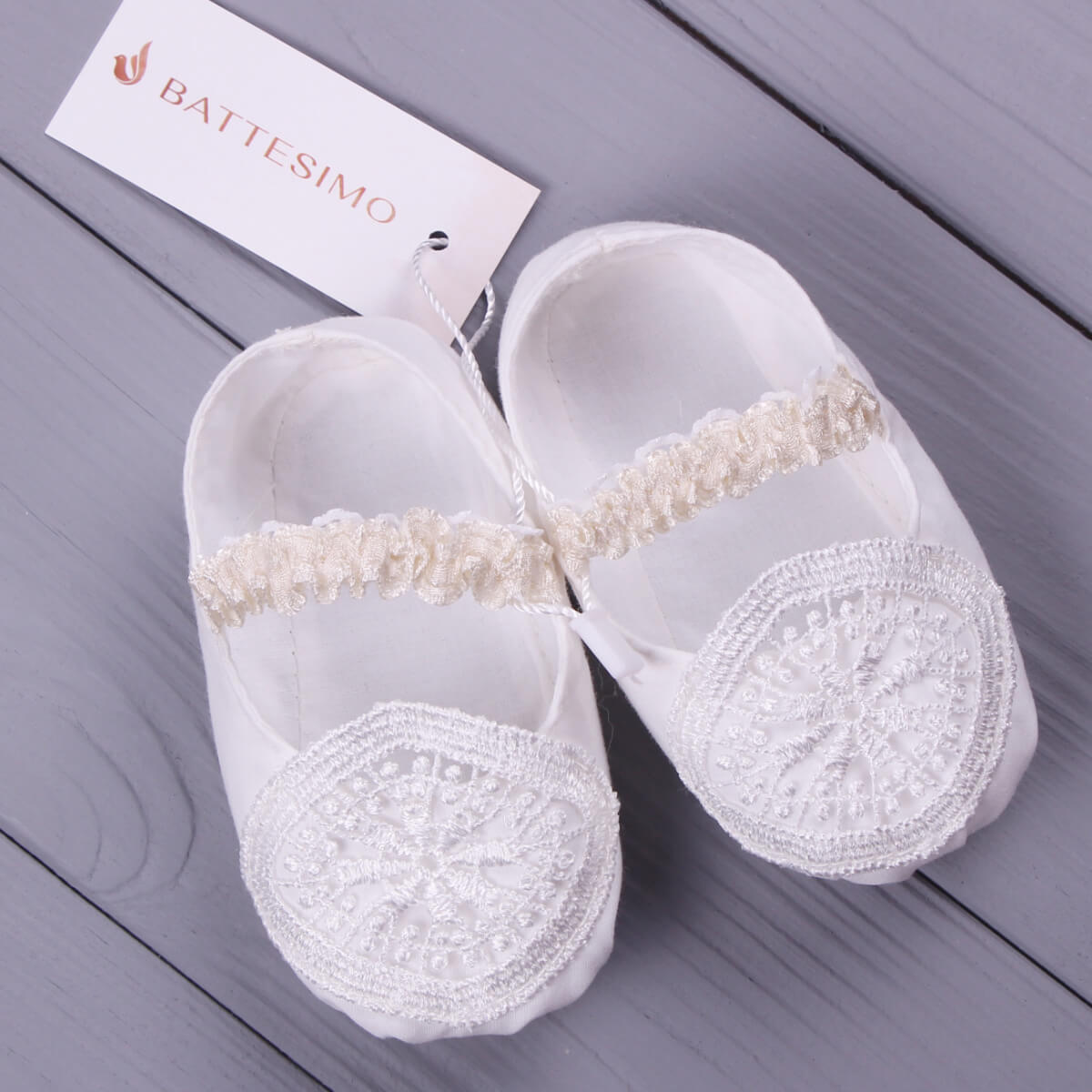  Baptism Booties with lace details on a gray wooden surface, featuring the brand 'Battesimo'.