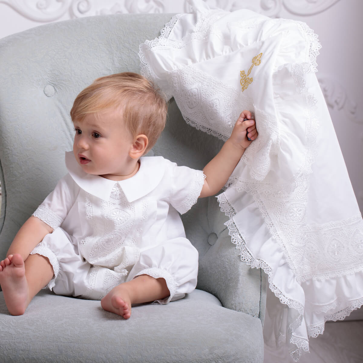 Baby in a white outfit holding a lacey white blanket on a gray chair.