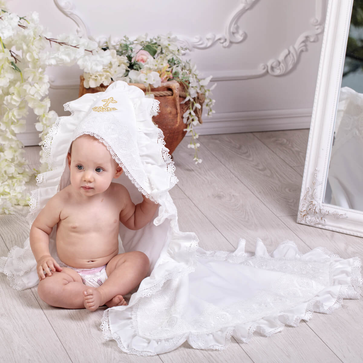 Baby sitting on a white lace blanket with a white lace headband, surrounded by flowers and a mirror.