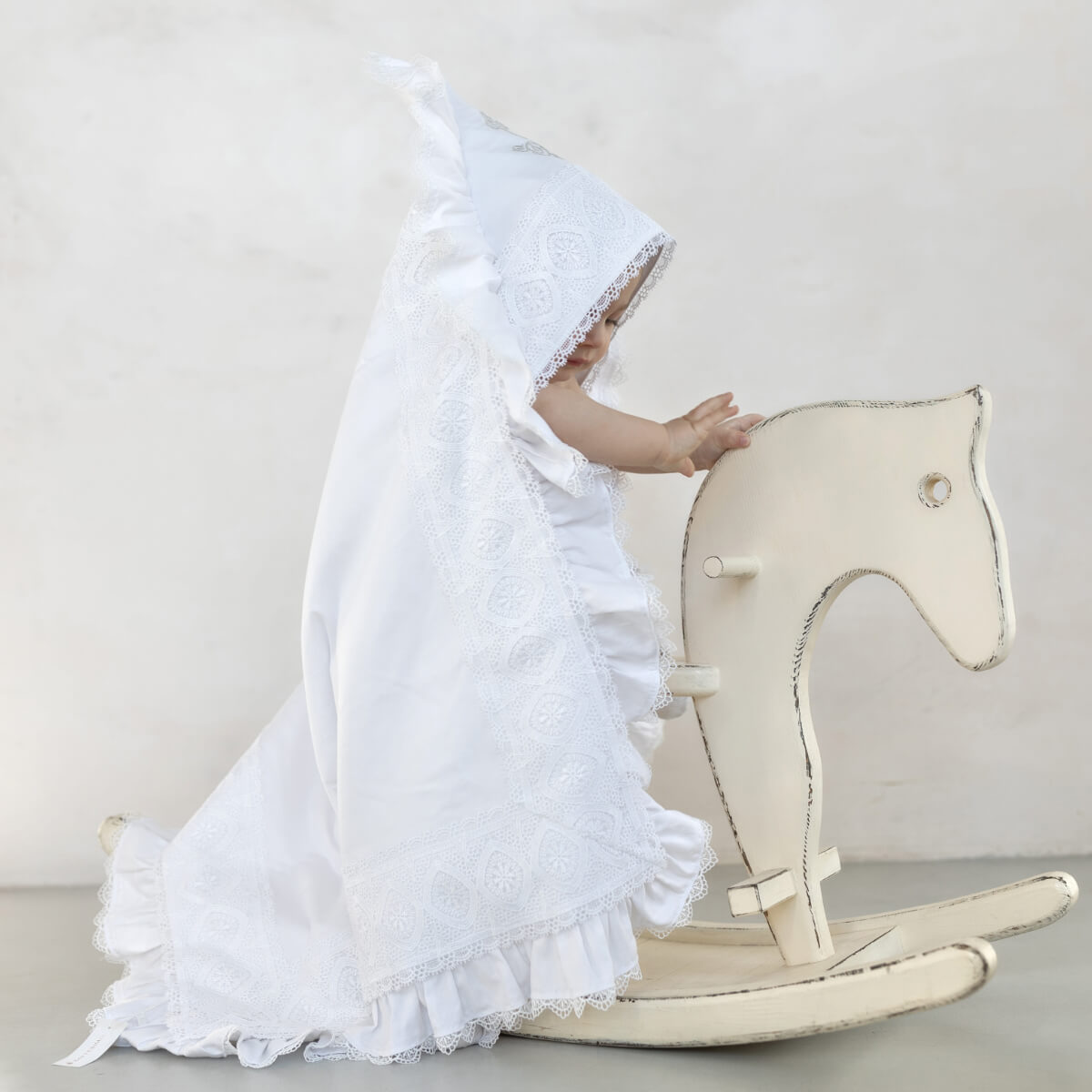 Child in a hooded baptism blanket playing with a rocking horse on a light gray background