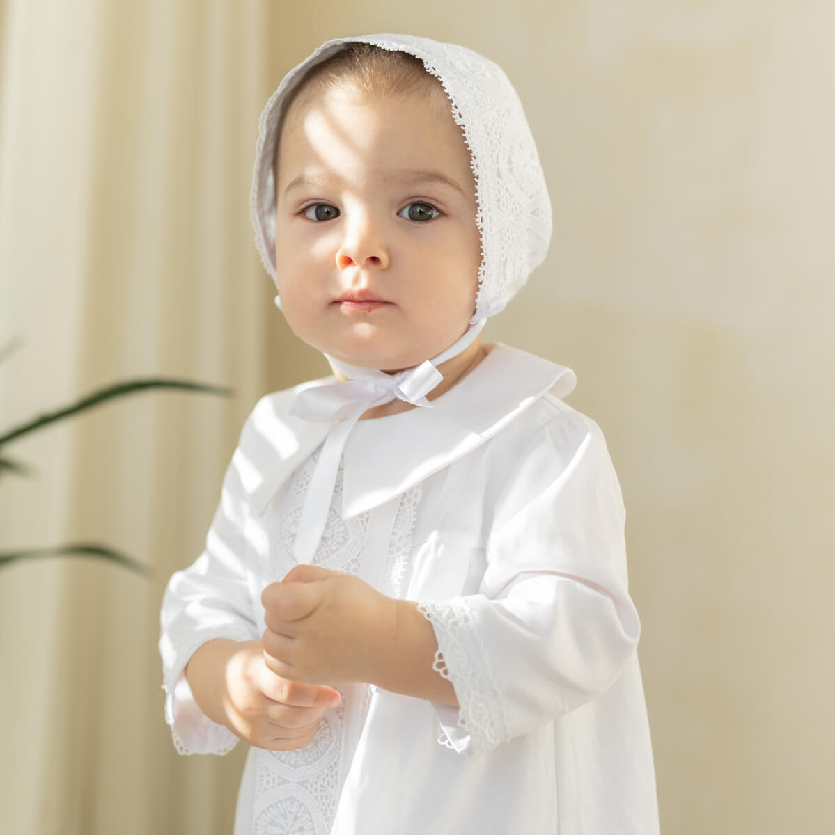 Baby wearing a white lace bonnet and christening gown against a beige background