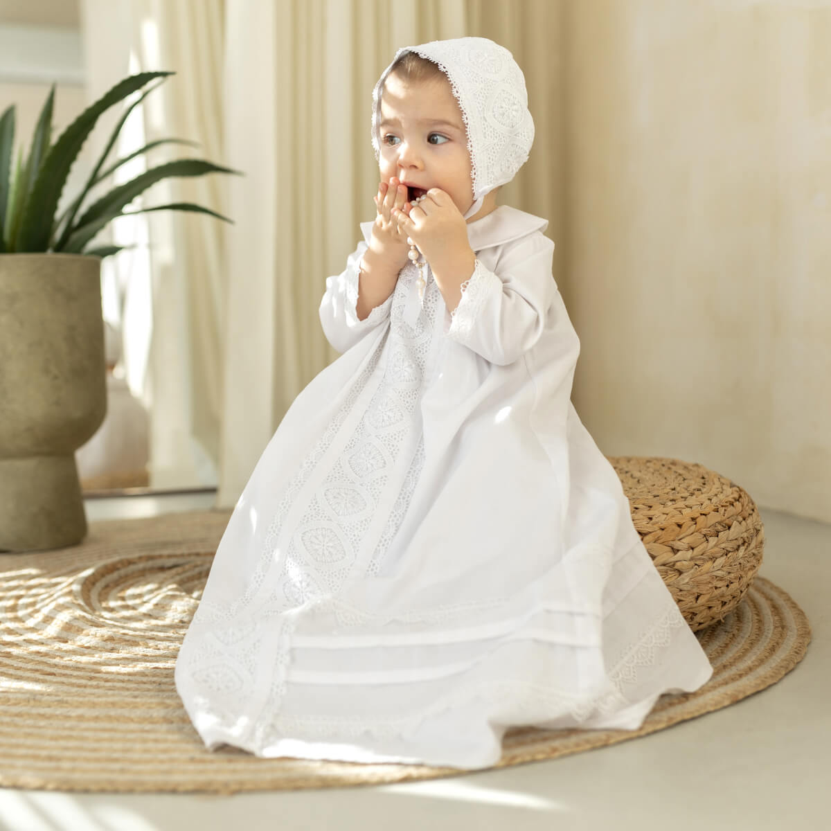 Baby in a white christening gown and bonnet sitting on a woven mat indoors.