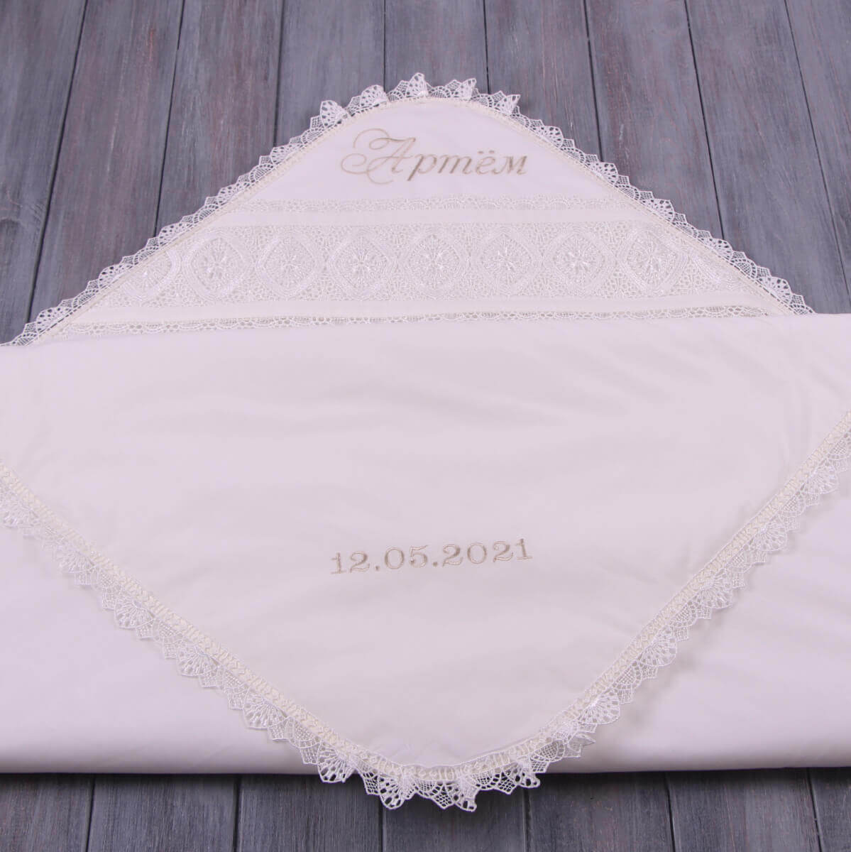 White lace-trimmed christening blanket with embroidered text on a wooden surface