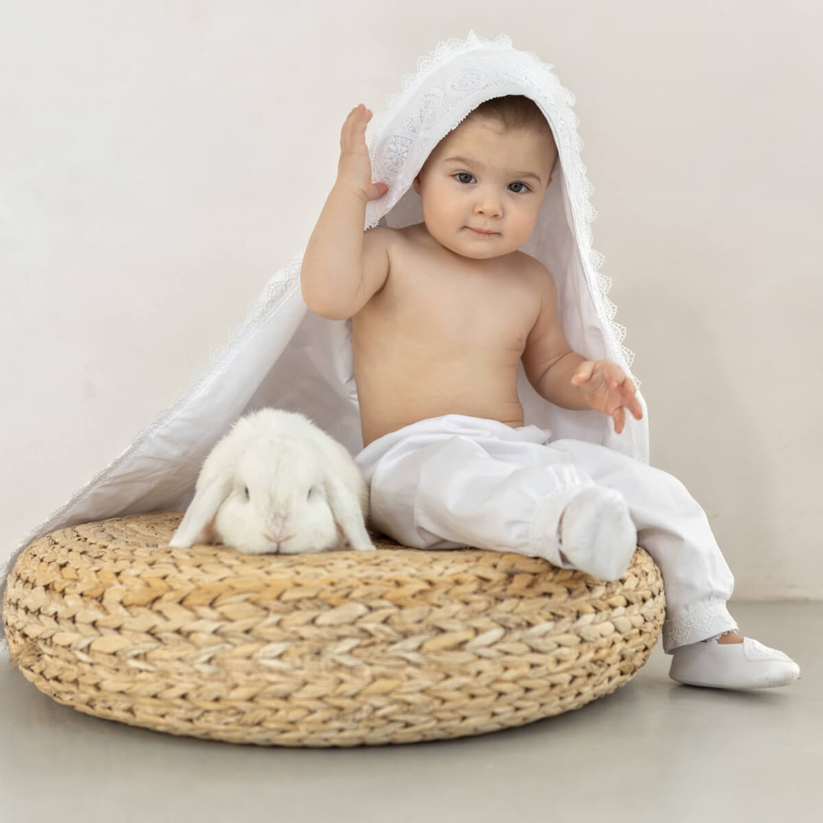Baby sitting on a woven stool with a white christening blanket 