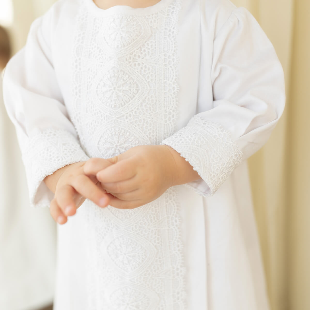 A close-up of a child wearing a white long-sleeve Boy Christening Gown  with intricate lace and embroidery details on the chest and cuffs, set against a soft beige background.