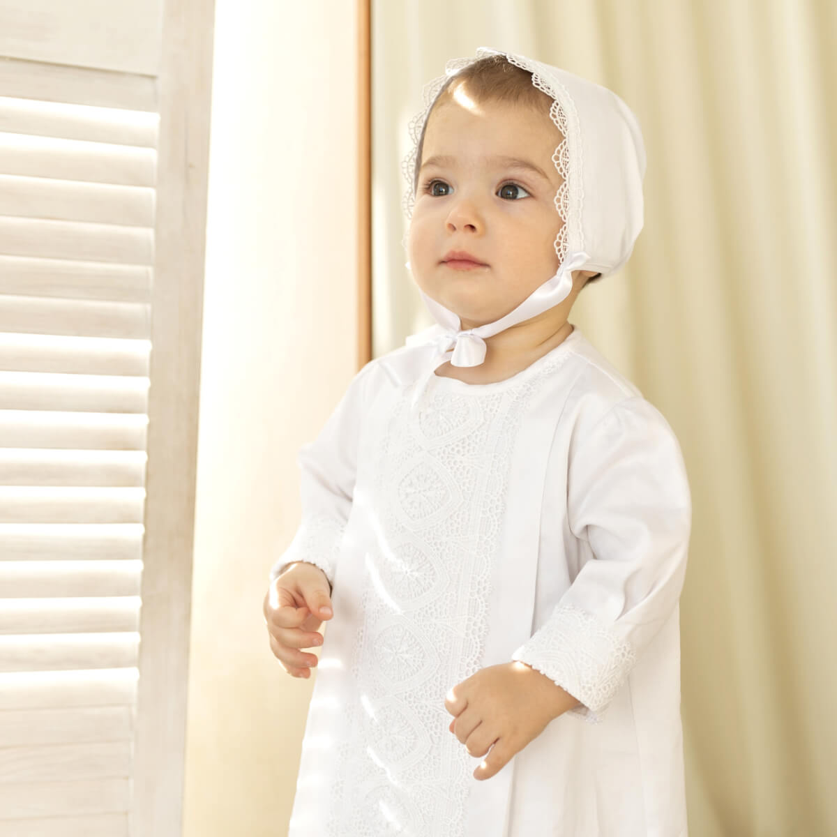 A baby wearing a white long-sleeve Boy Christening Gown and matching lace-trim bonnet, standing indoors near a window with soft warm light.