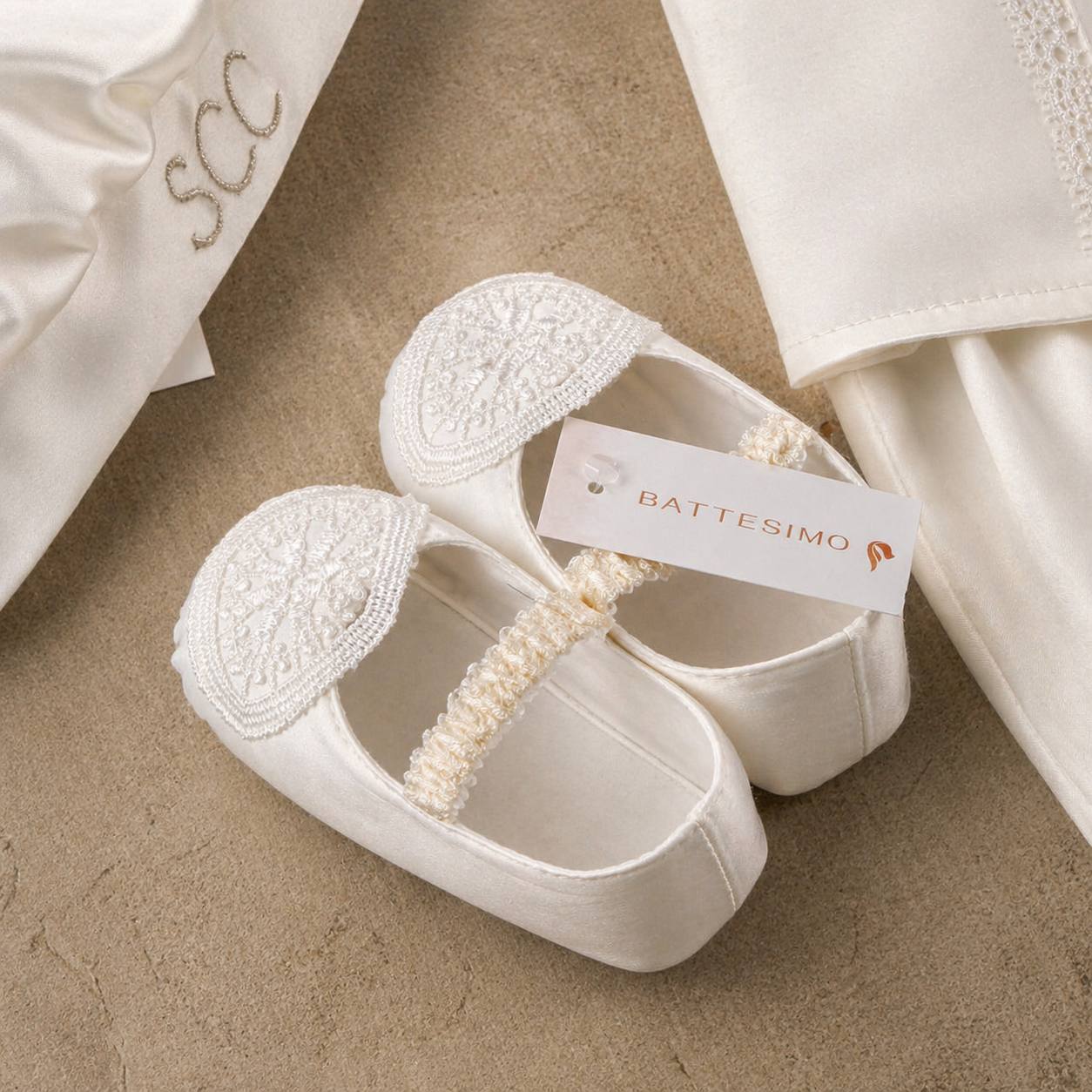 White baby shoes with lace details and a headband on a beige surface, featuring a Battesimo label.
