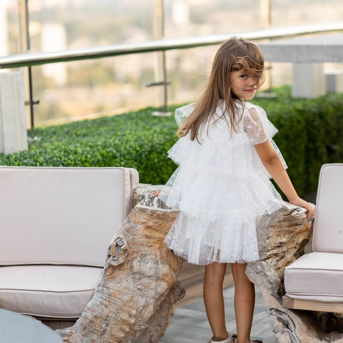 Young girl in a white dress standing outdoors near a stone and chair.