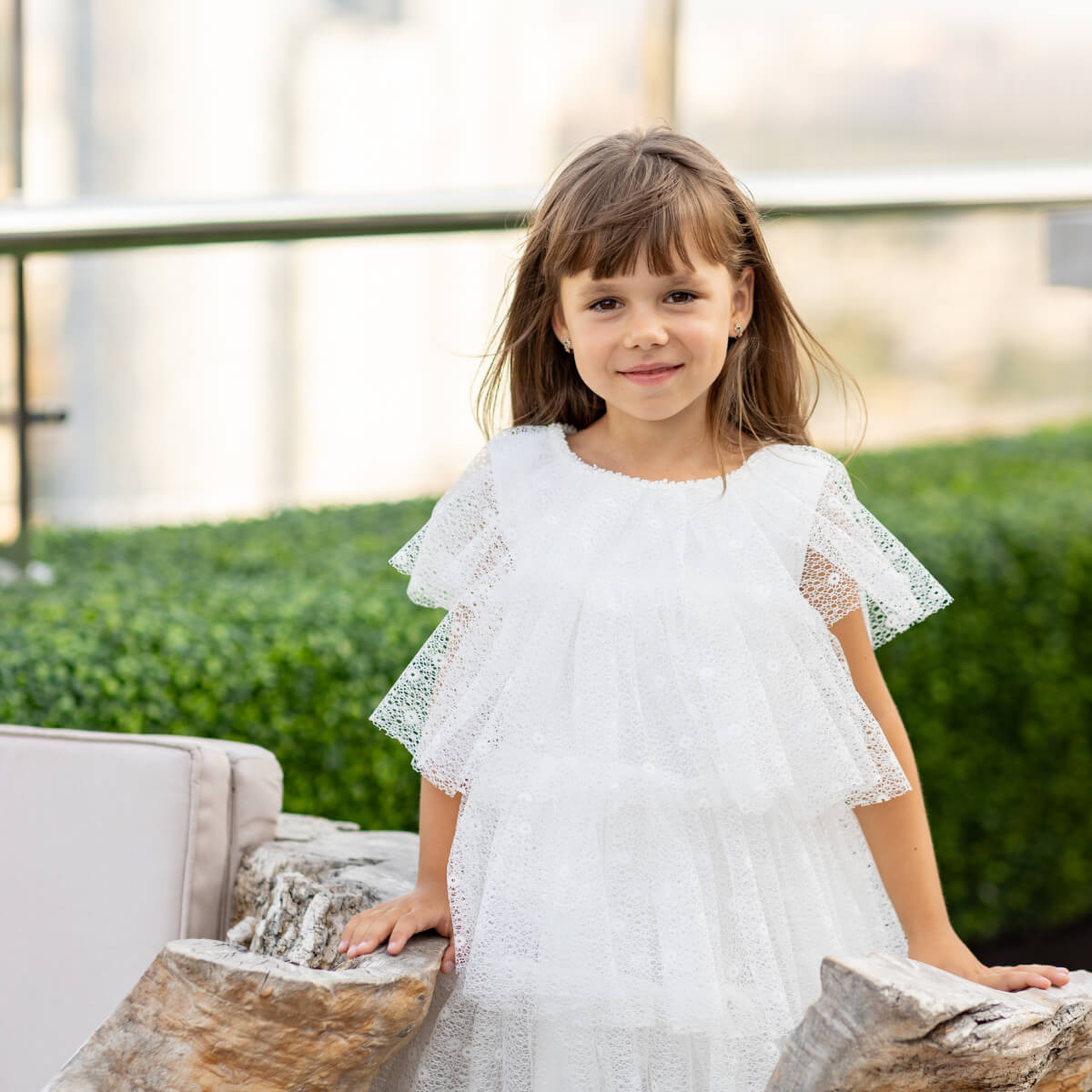Young girl in a white dress sitting on a stone bench with greenery in the background