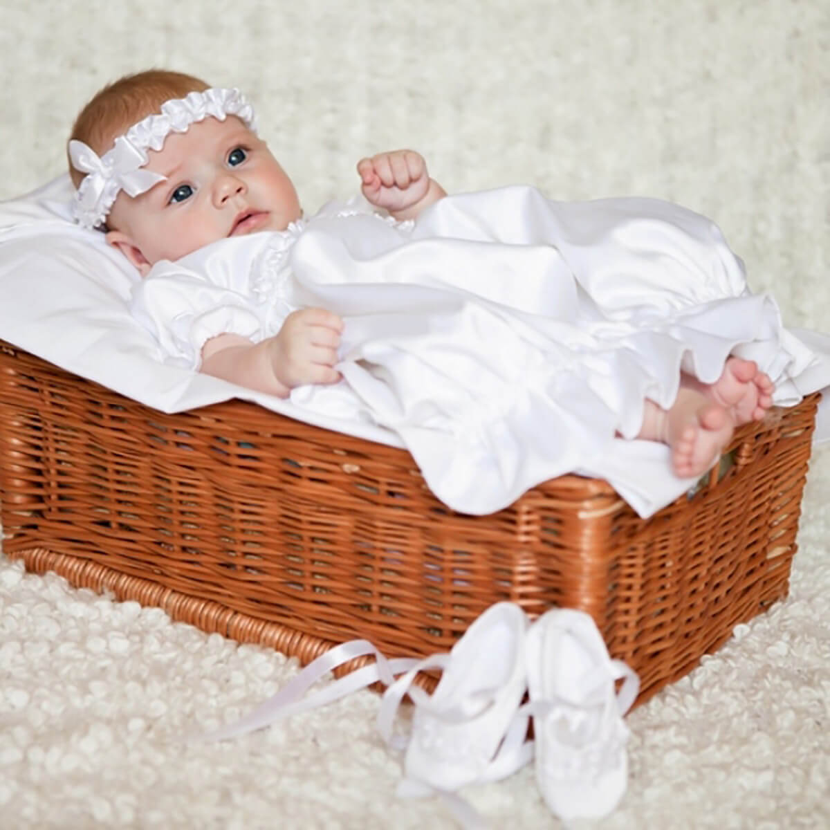 Baby in a Newborn Christening Gown and headband lying in a wicker basket on a soft surface.