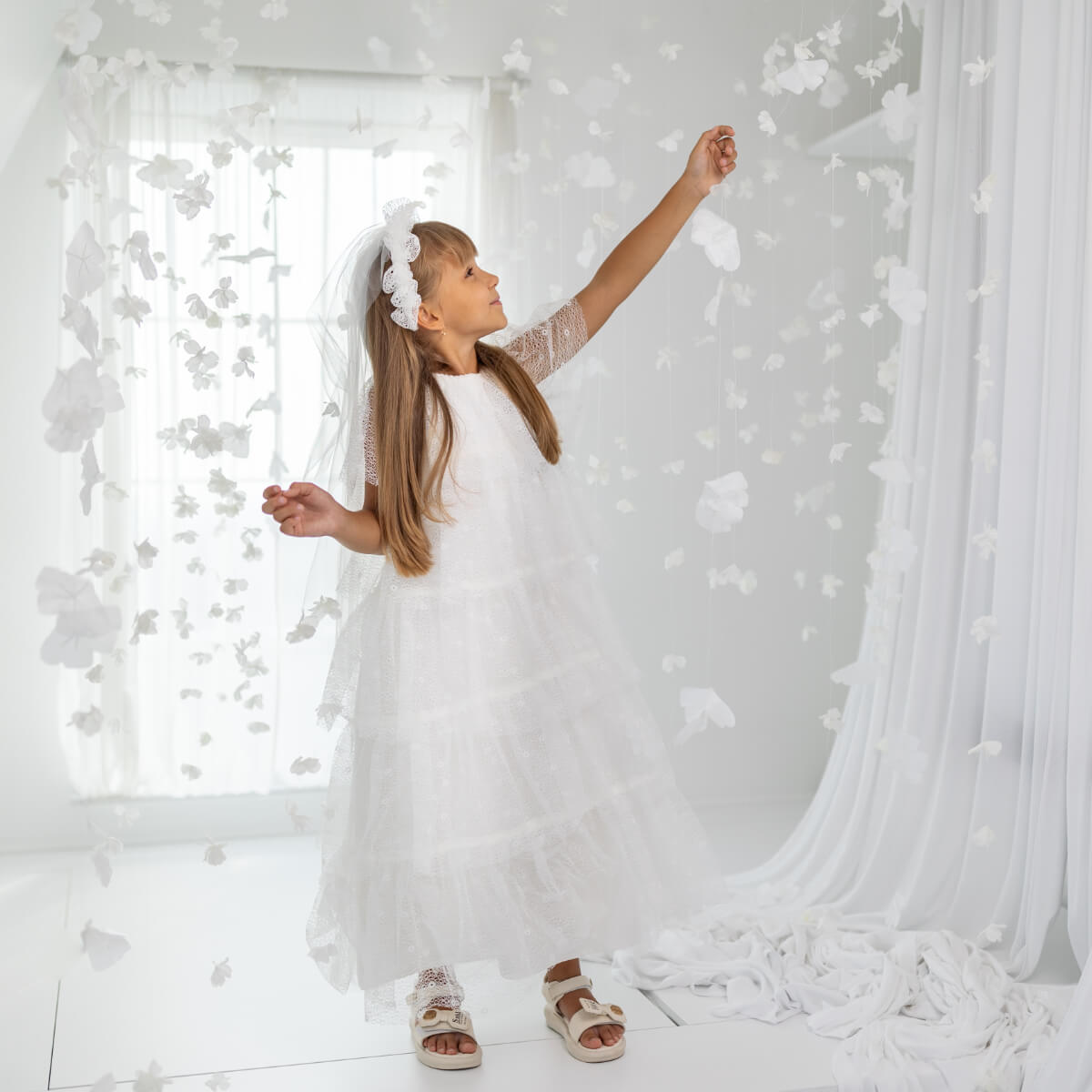 Young girl in Holy First Communion standing in a room with white curtains and decor.