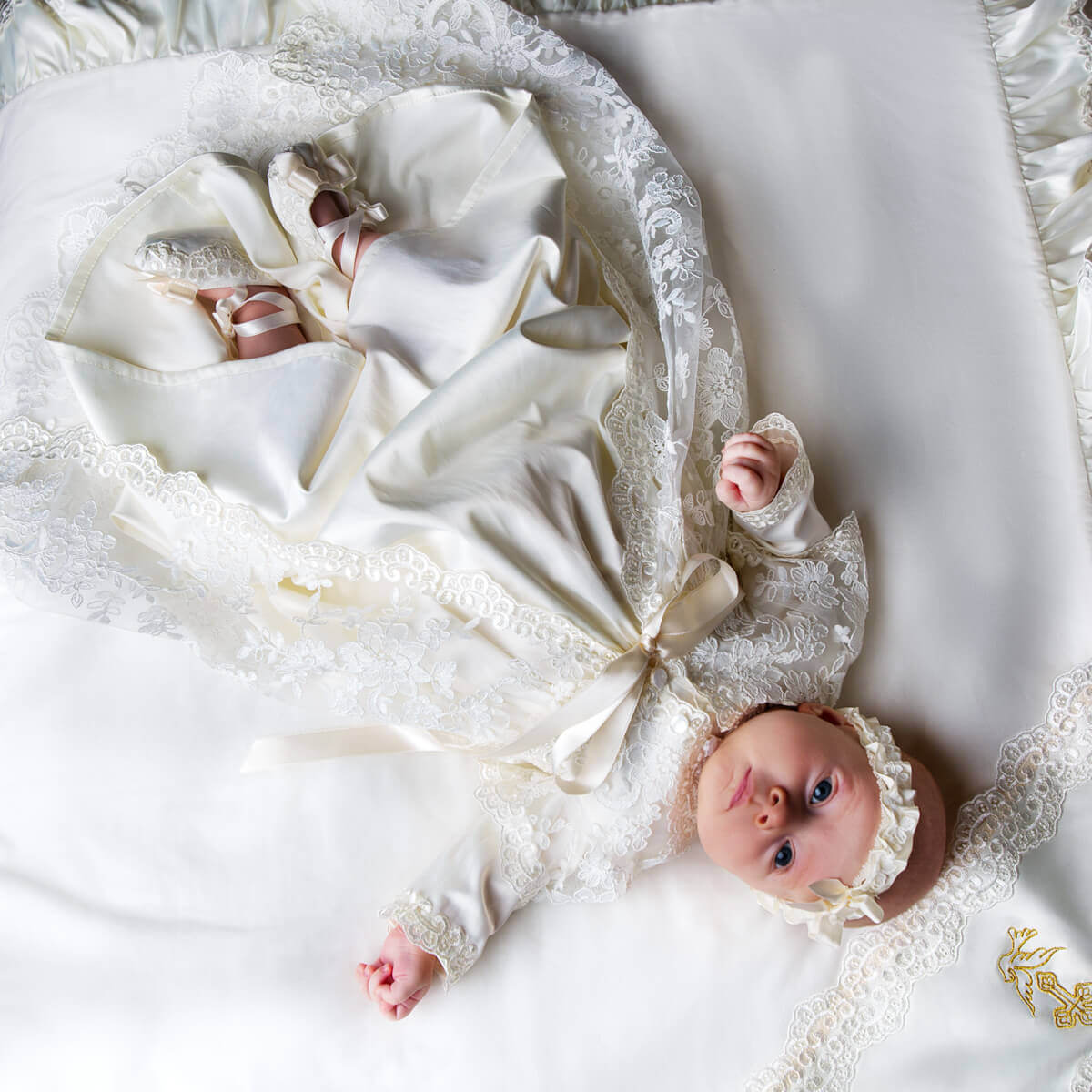 Baby in a  Christening Gown for Girls lying on a white surface with another baby peeking out from a white crib.