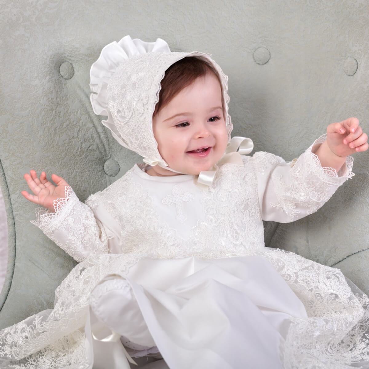 Baby in a Heirloom Christening Gown for Girls and bonnet sitting on a gray couch.