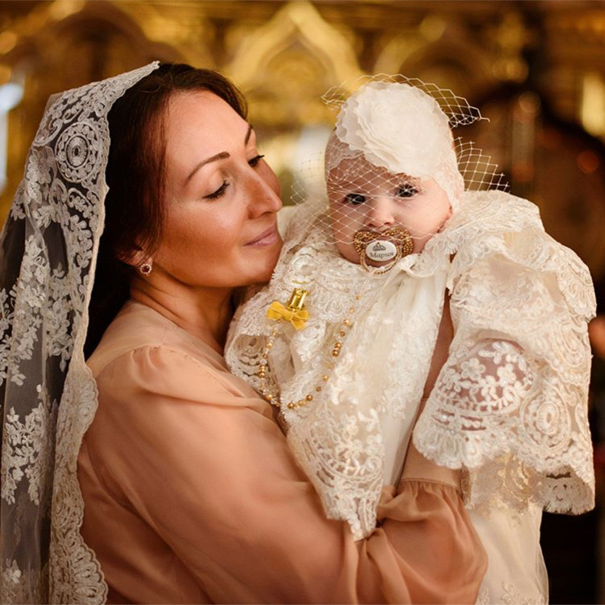 Woman  in Catholic Church Mantilla holding a baby dressed in white lace attire with a blurred golden background