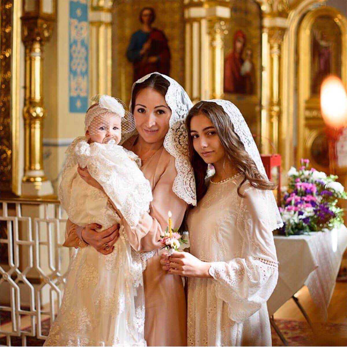Two women in white dresses with Catholic Church Mantillas holding a baby in a church setting.