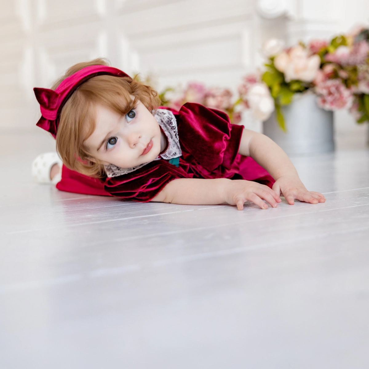 Child in a red dress with a large bow sitting on a white surface with flowers in the background