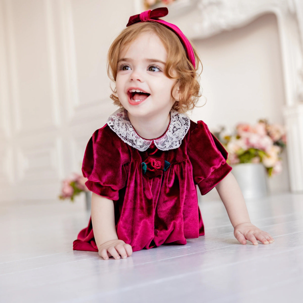 Child wearing a red velvet dress with lace collar and flower detail, sitting on a white surface.