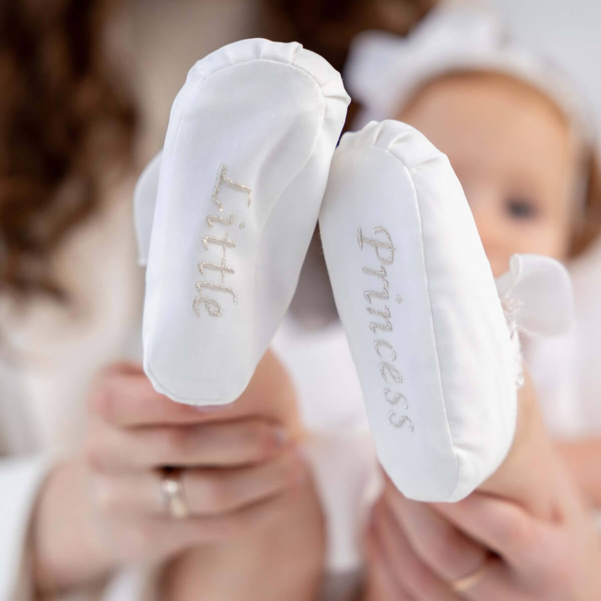 White booties with 'Little Princess' text held by a person, with a baby in the background.