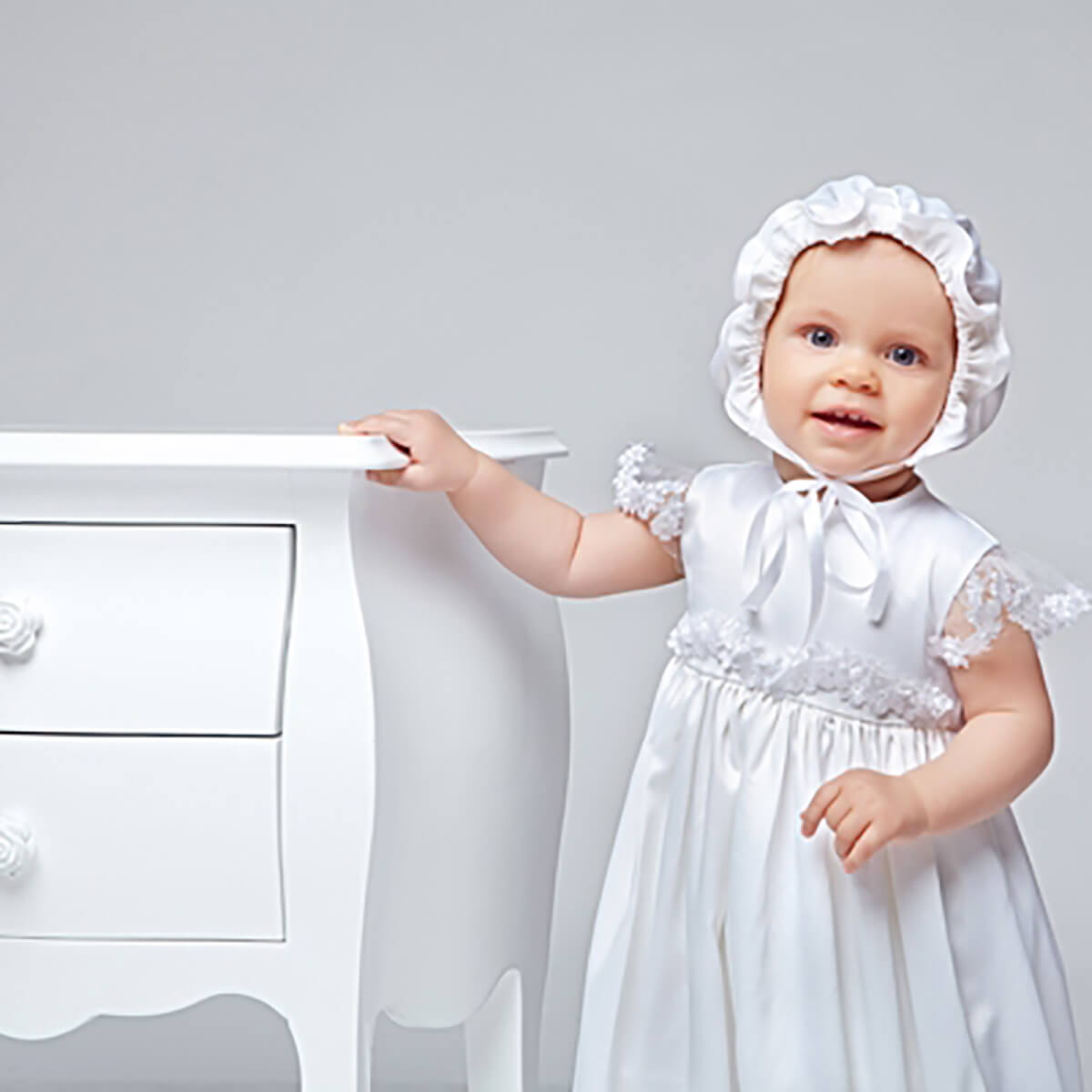 Baby in a Christening Gown for Girls and bonnet standing next to a white dresser.