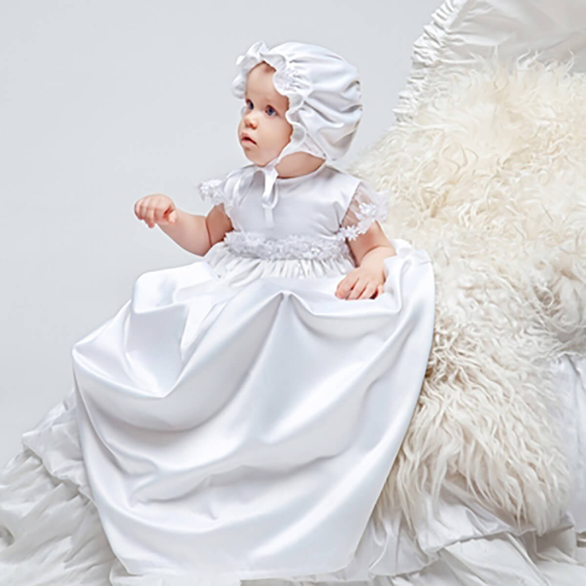 Baby in a  Christening Gown for Girls and bonnet sitting on a fluffy white rug.