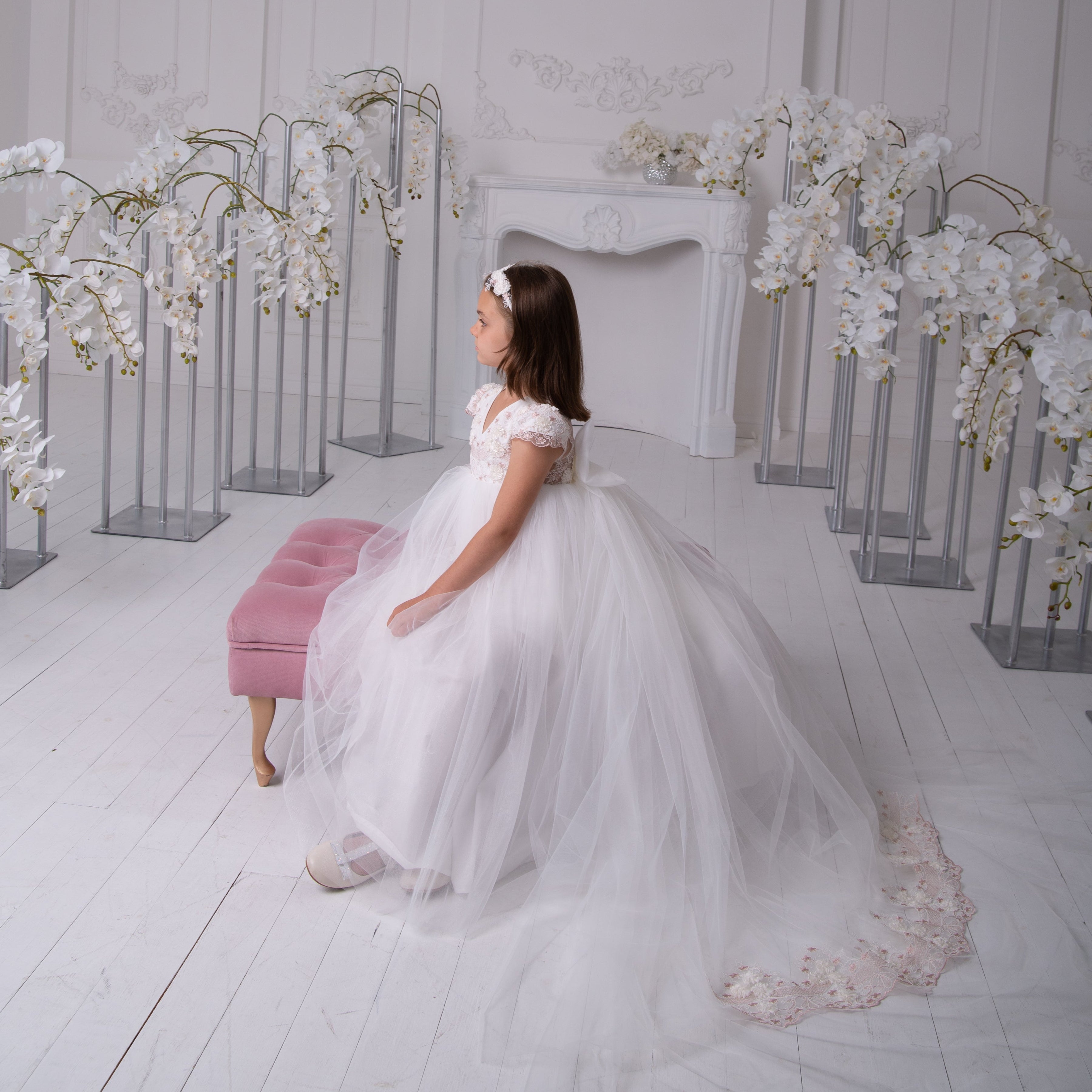 Young girl in a white dress standing in a room with floral decorations and a chandelier.