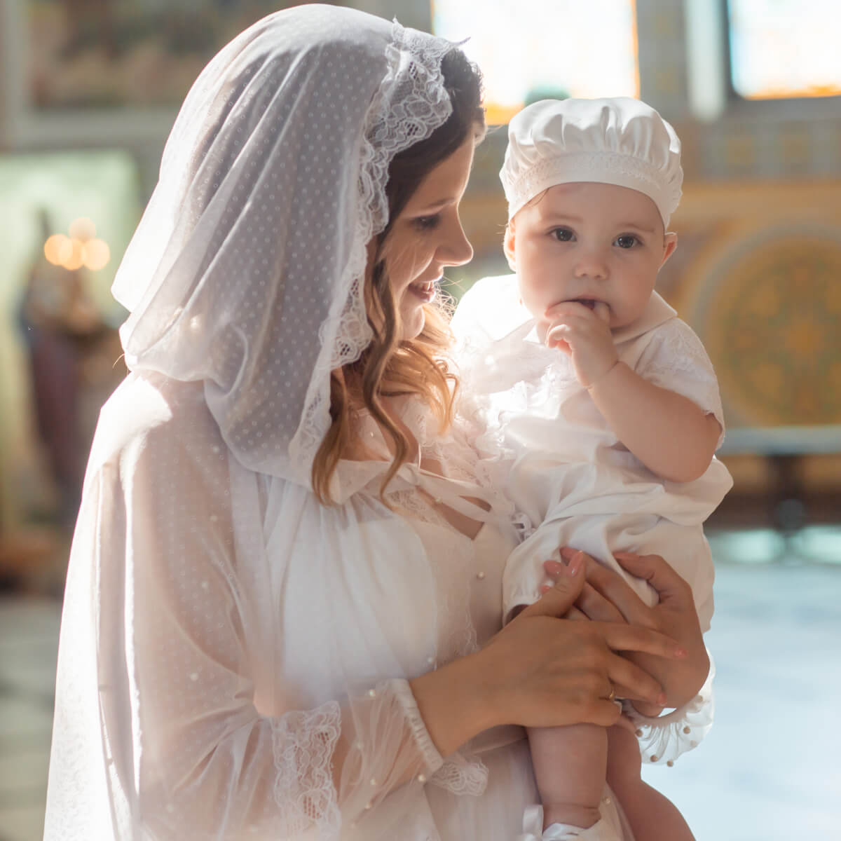 Woman in white dress holding a baby in Boy Christening Hat