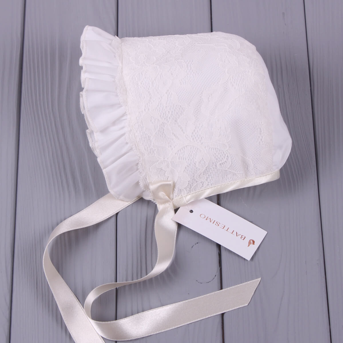 Christening Bonnet with lace details on a gray wooden surface, featuring a brand tag.