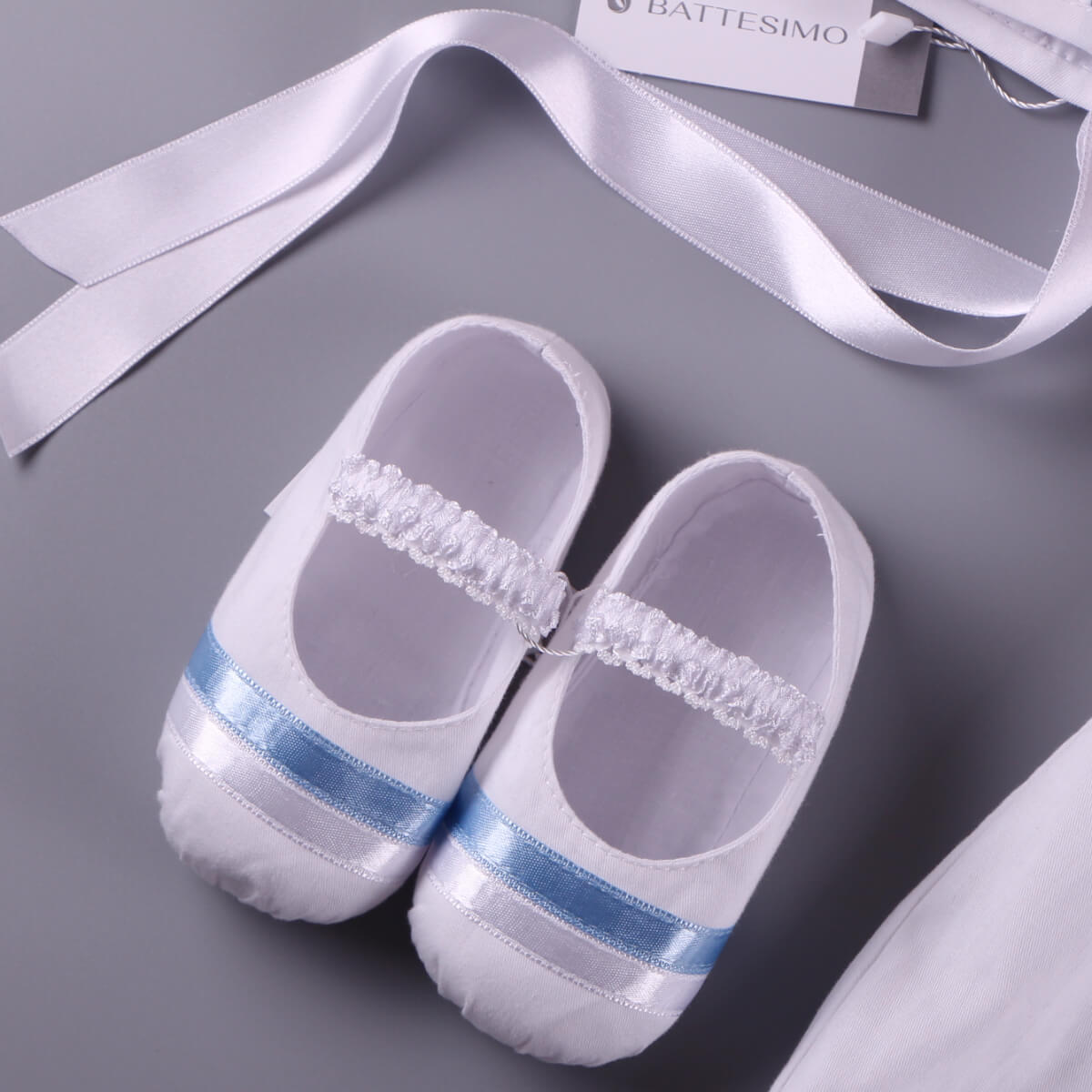 White baby shoes with blue stripes and decorative ribbons , as a pert of Christening Outfit on a gray background