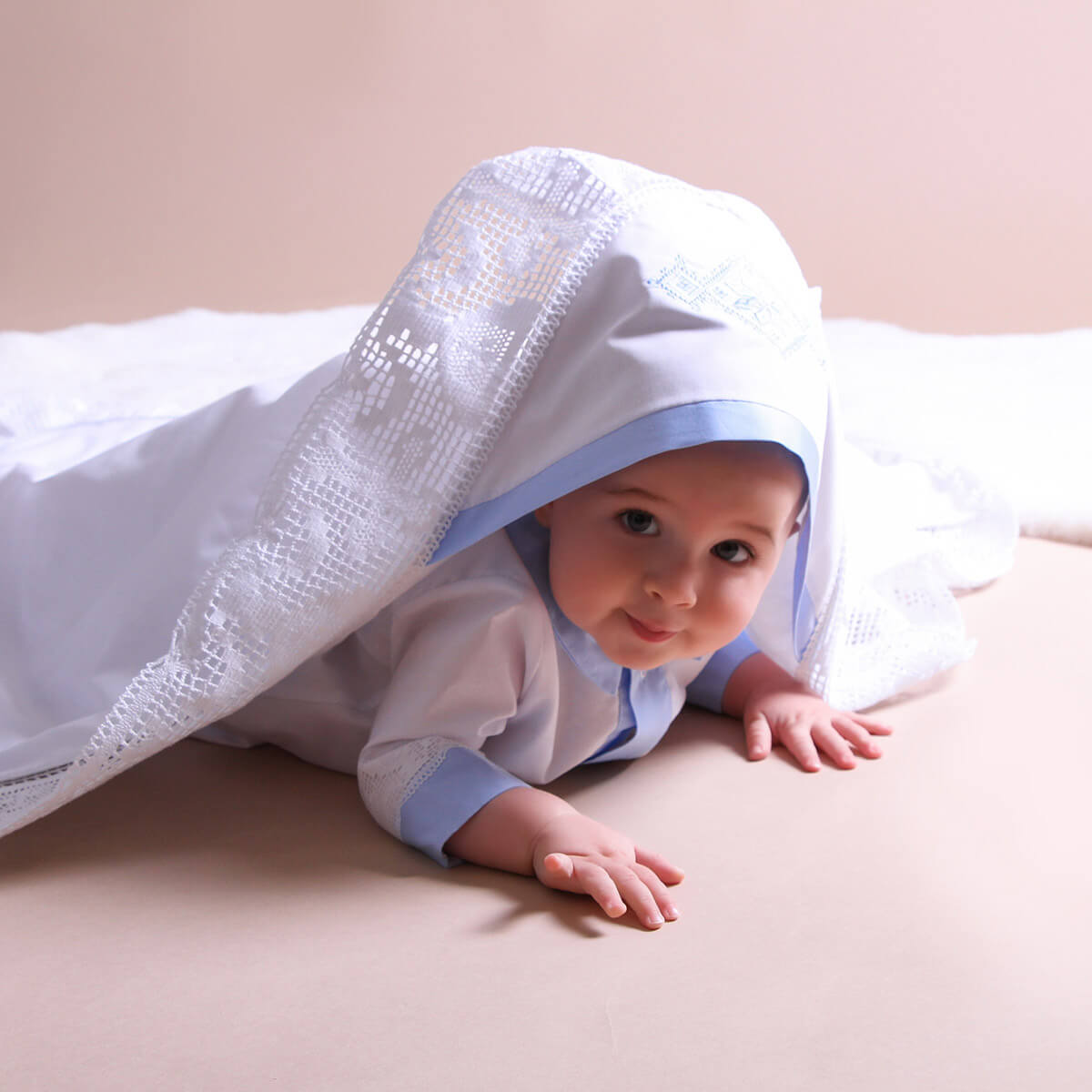 Baby wearing a white outfit with blue trim peeking out from under a white blanket on a beige surface.