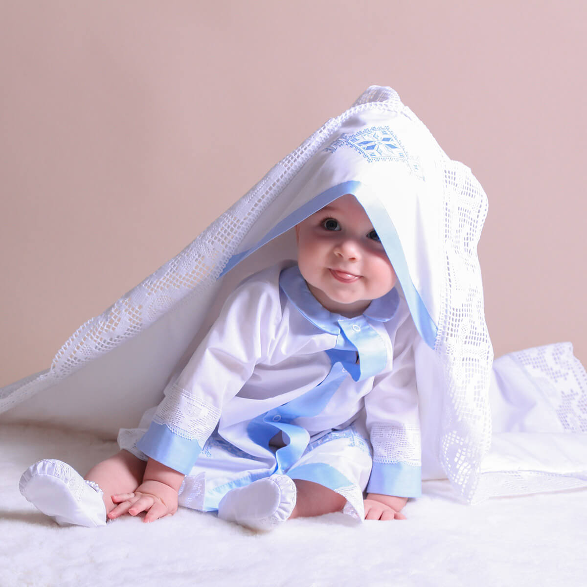 Baby in a white outfit peeking from under a white christening blanket 