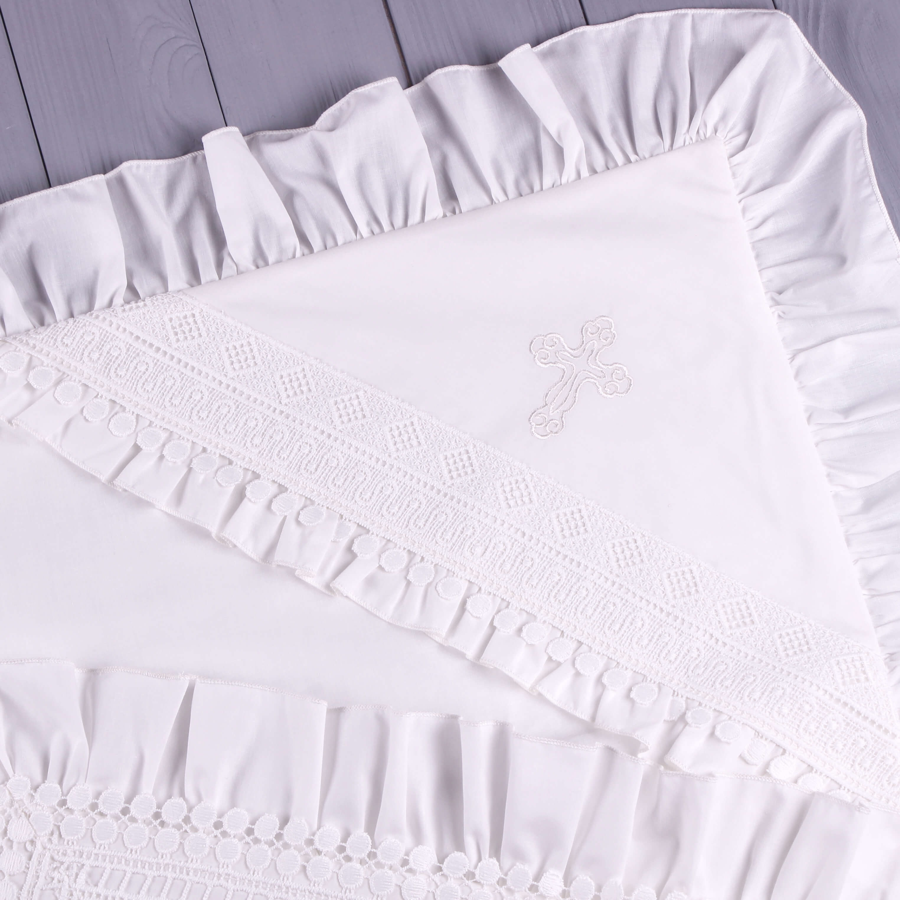 White embroidered cross on a white baptism blanket with ruffled edges.