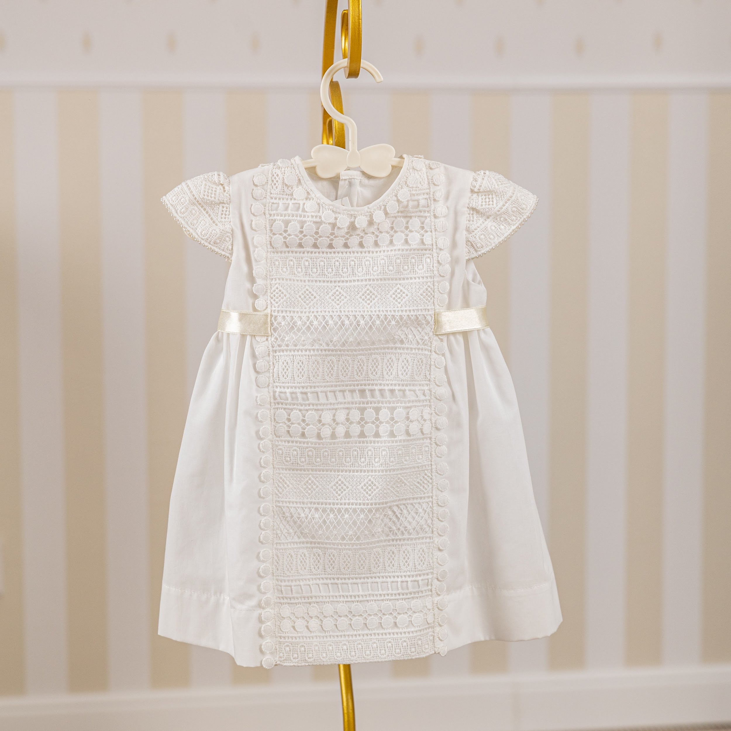 Baptism Dress with Macramé Lace on a hanger against a striped background