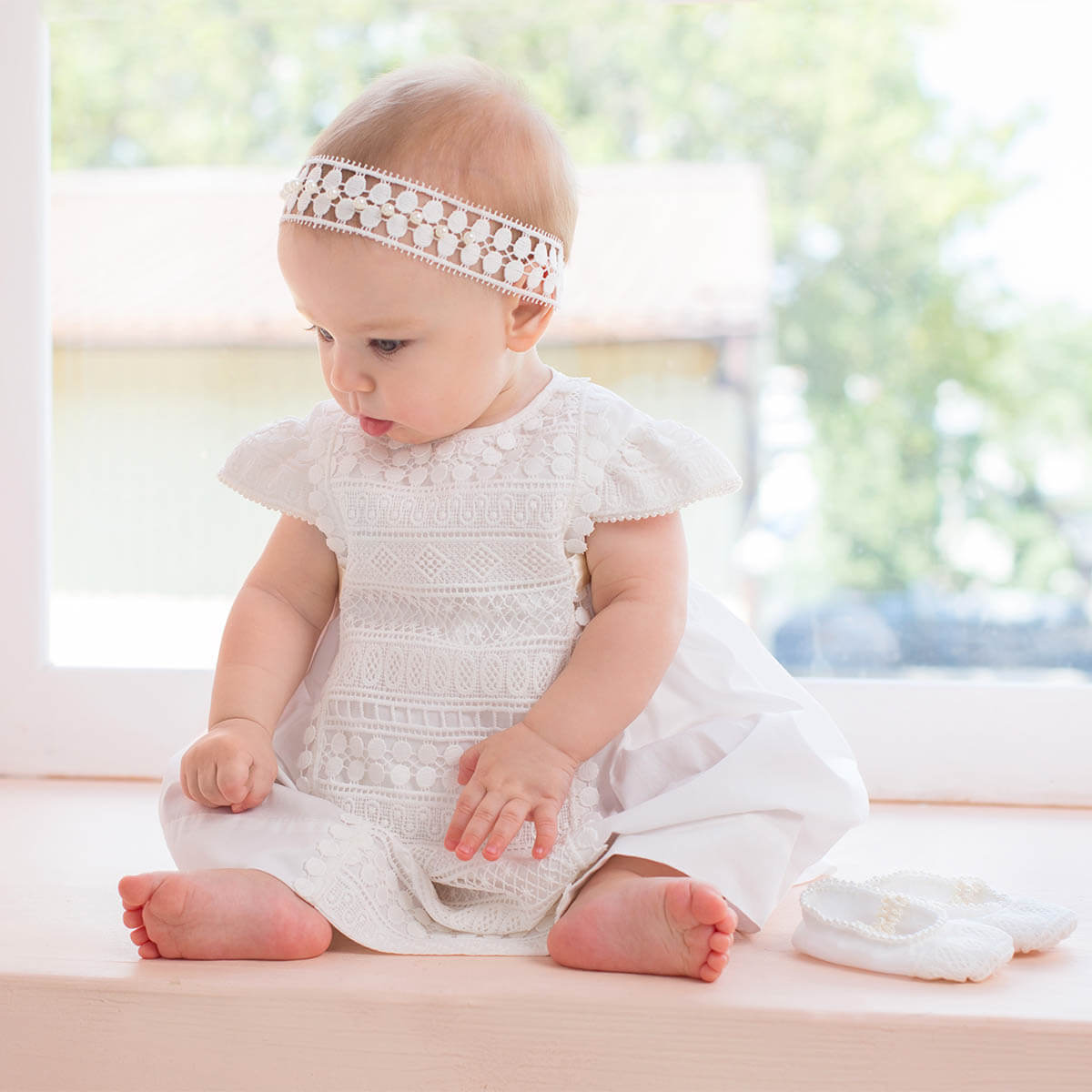 Baby in a Baptism Dress with Macramé Lace sitting on a light surface with a blurred natural background