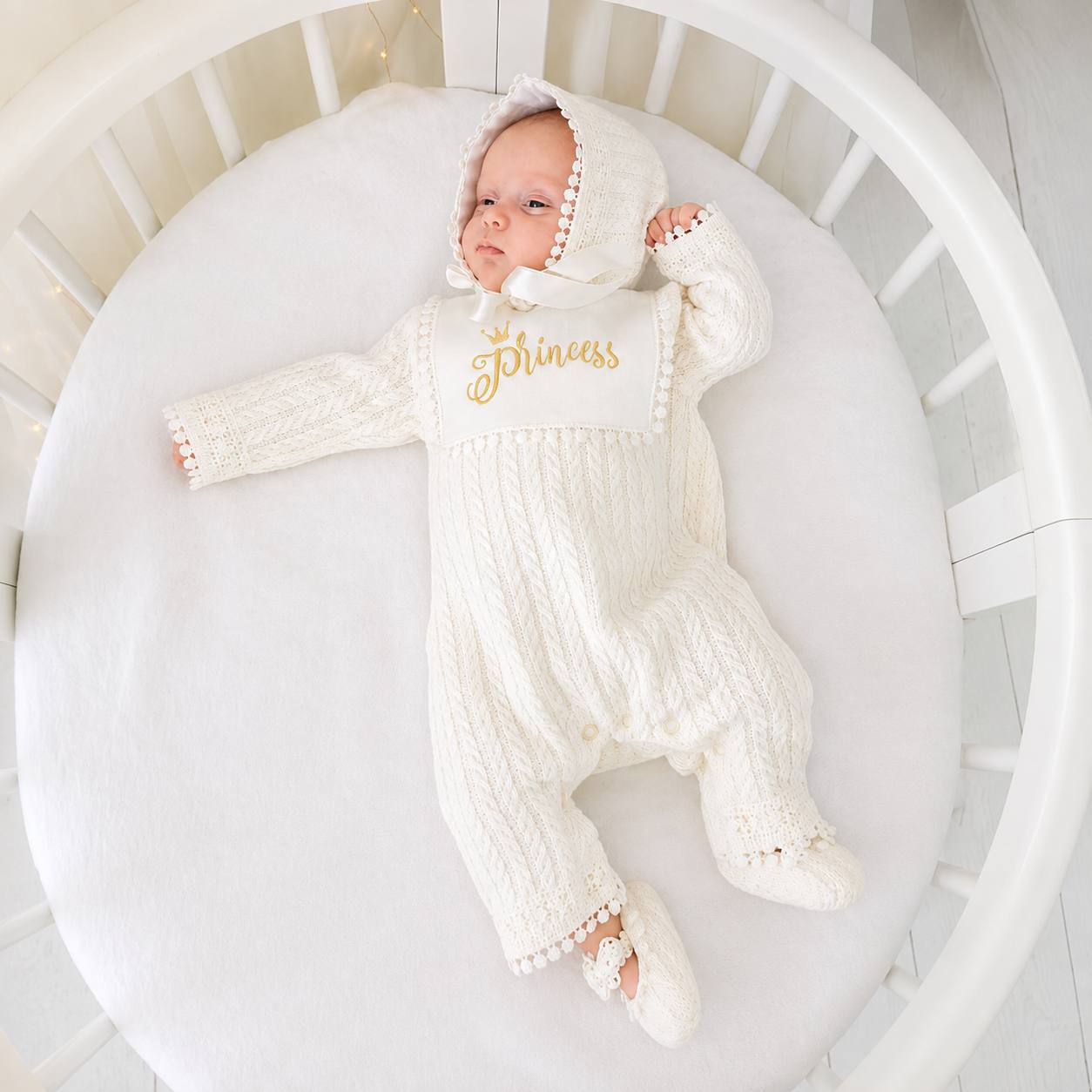 A baby lying on a white cushion inside a white round bassinet, wearing an off-white cable-knit hooded footed romper with gold 'Princess' lettering on the chest and matching knit booties.