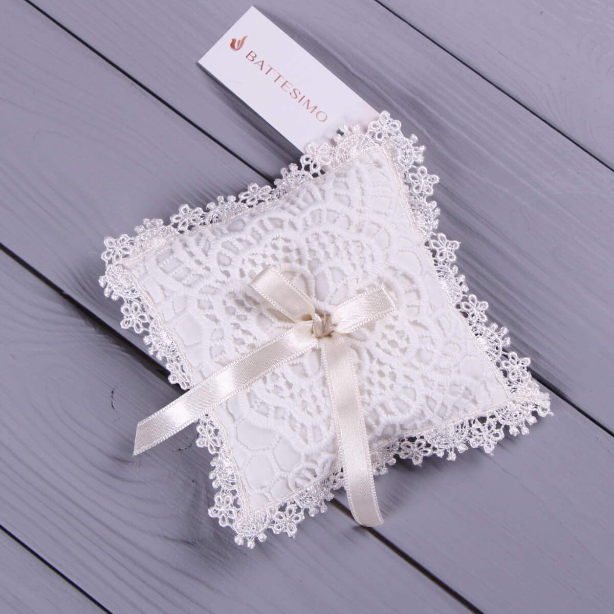 Christening Cross Pillow with a ribbon on a wooden surface, branded 'Battesimo'.