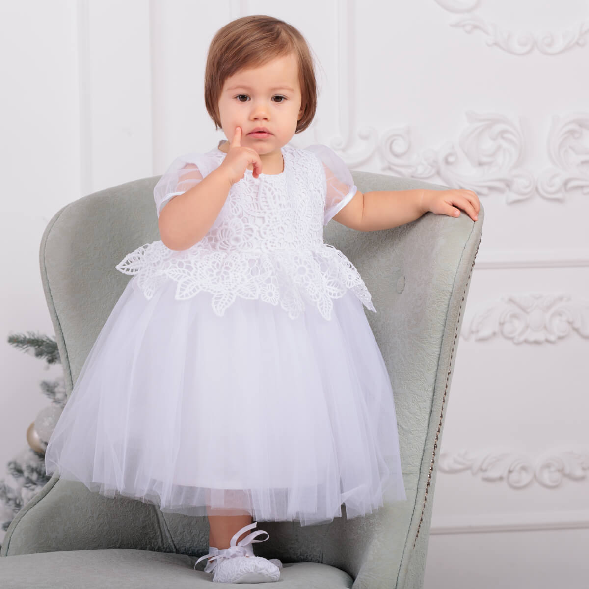 Child wearing a Christening Lace Dress sitting on a gray chair against a white wall.