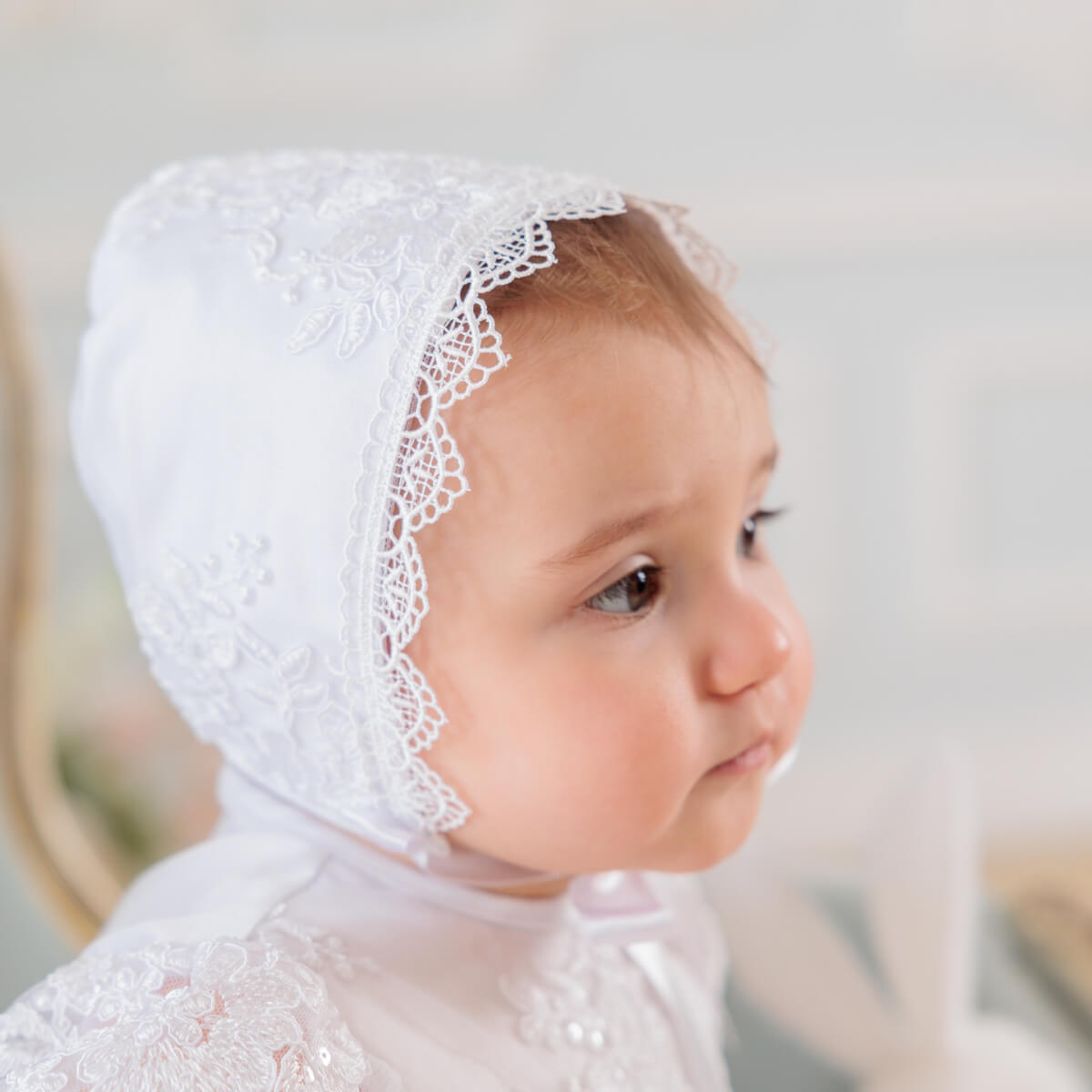 Baby wearing a white lace bonnet of  Baptism Set for Girl against a blurred background