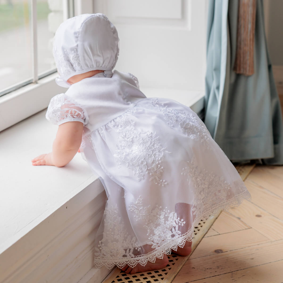 Baby in a  Baptism Set for Girl  and bonnet standing on a windowsill.
