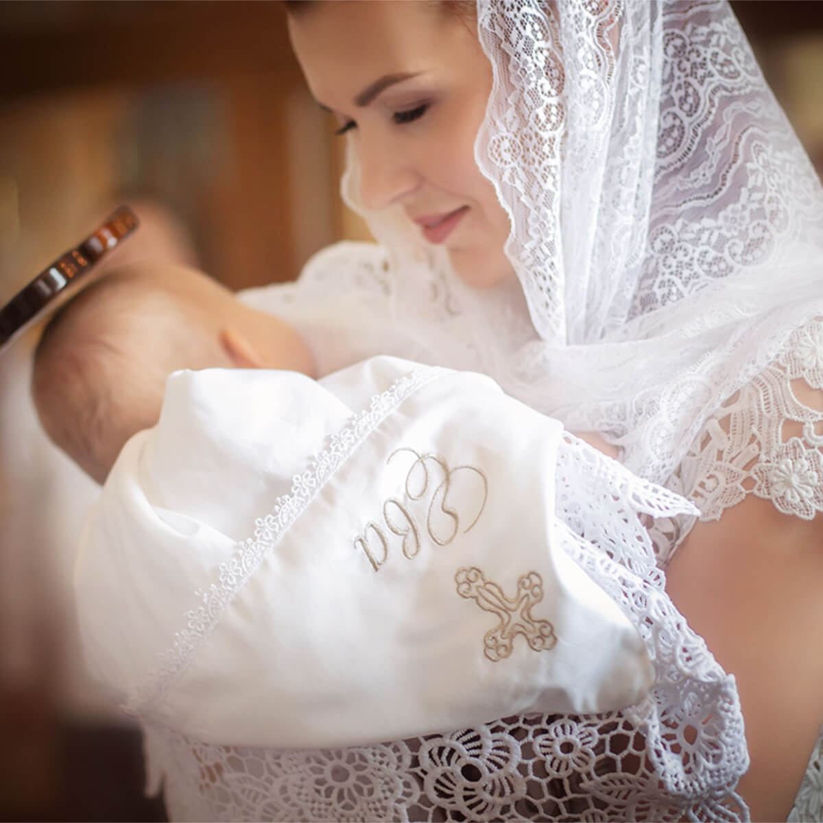 Woman holding a baby wrapped in a white christening blanket with embroidered initials and cross, wearing a white lace headscarf.