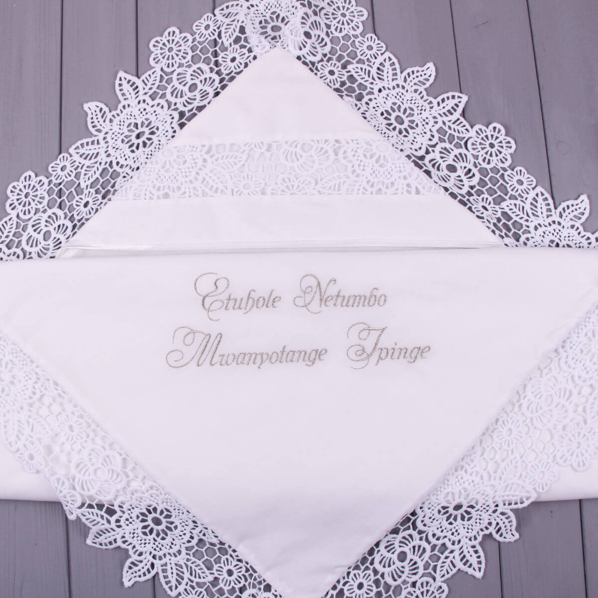 White lace-trimmed christening blanket with embroidered text on a wooden surface