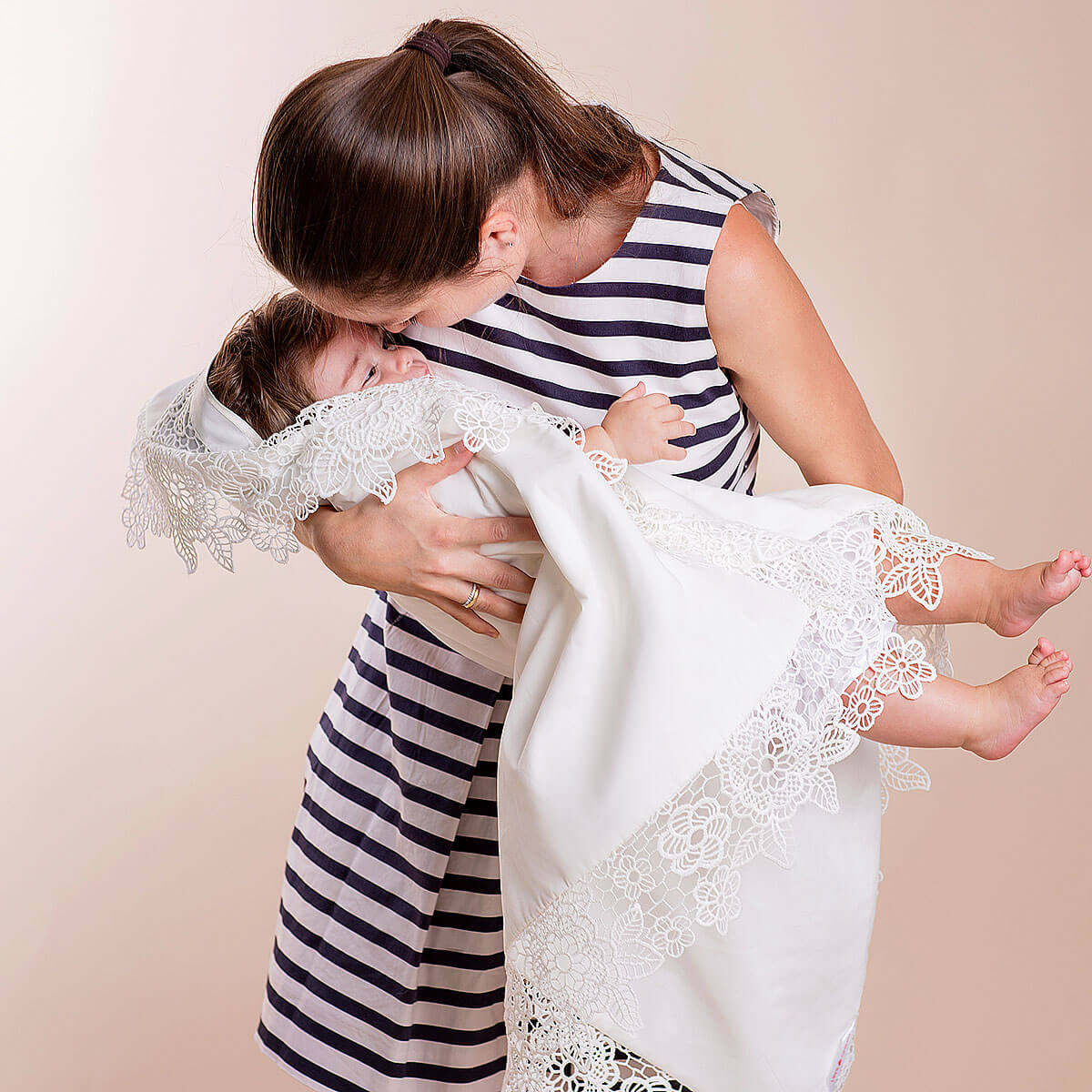 Woman holding a baby wrapped in a white blanket with lace trim against a beige background