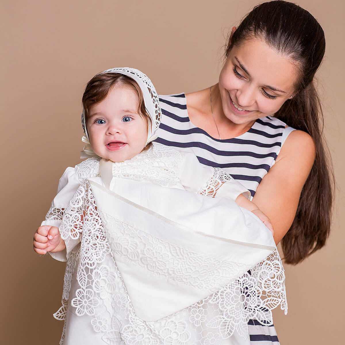 Woman holding a baby wrapped in a white lace blanket against a brown background