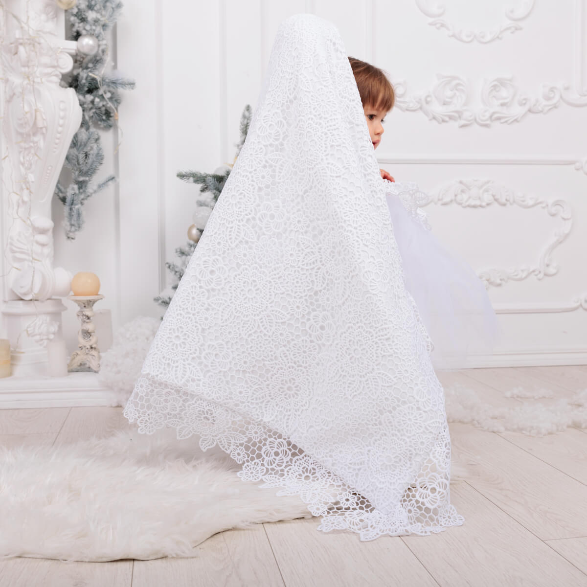 Child peeking out from under a large white lace blanket in a decorated room.